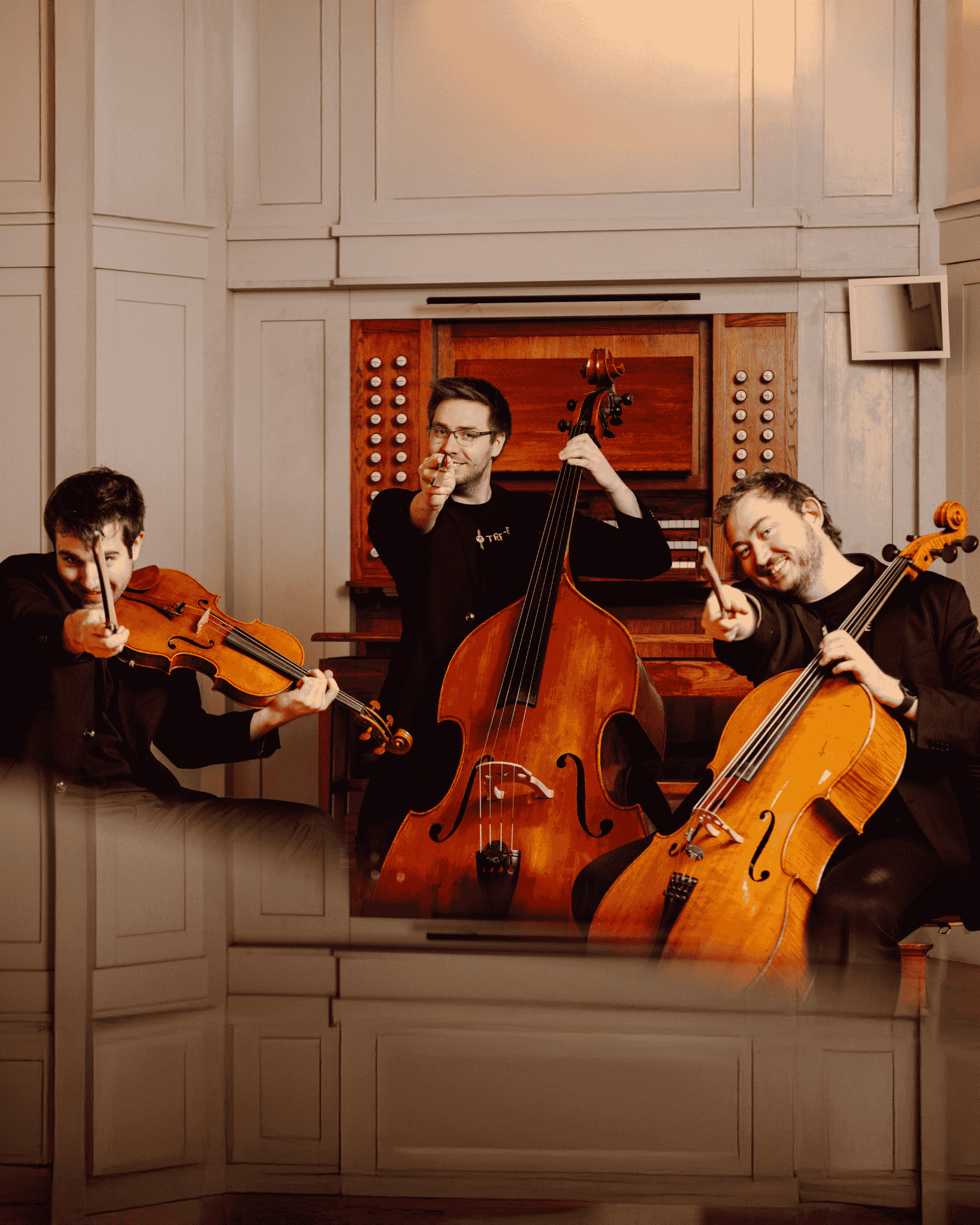 Tre styrkemusikere på bratsj, kontraball og cello sitter på stoler i en halvsirkel med instrumentene plasser til å spille og peker mot kameraet med buen